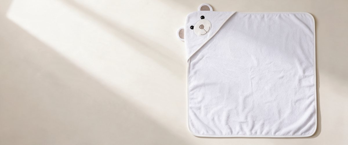 The Bear hooded towel laid flat on a cream surface in soft window light, showing its full 90 × 90 cm size